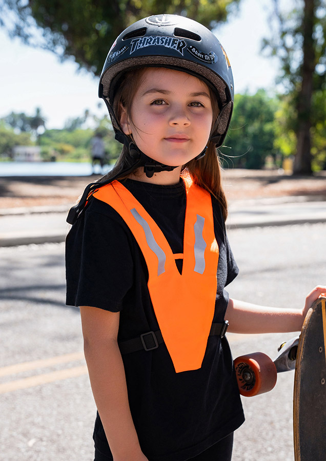 A child wearing an orange Korntex multifunctional collar with reflective stripes is standing in a park.