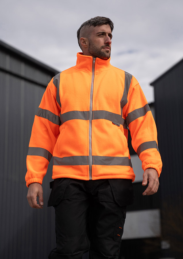 Adult man wearing a signal orange Korntex hi-vis fleece jacket with reflective stripes walking in front of an industrial buil