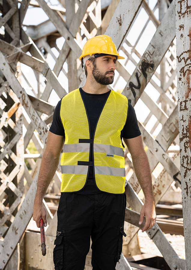Adult construction worker stands on a building site in a yellow Korntex mesh safety vest and yellow helmet holding tools