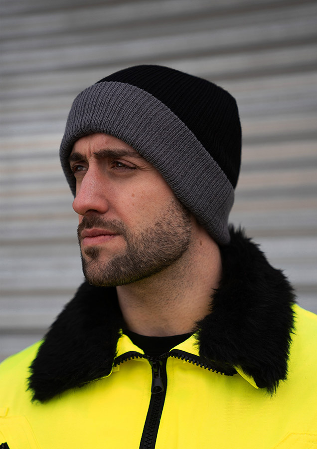 An adult wears an embroidered, black and grey Korntex knitted hat in front of a grey wall