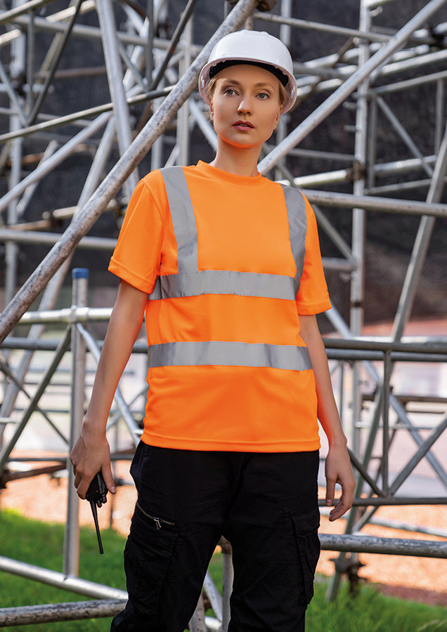 A woman, wearing a signal orange Korntex high-visibility T-shirt and a white hard hat, stands in front of scaffolding at an industrial construction site.