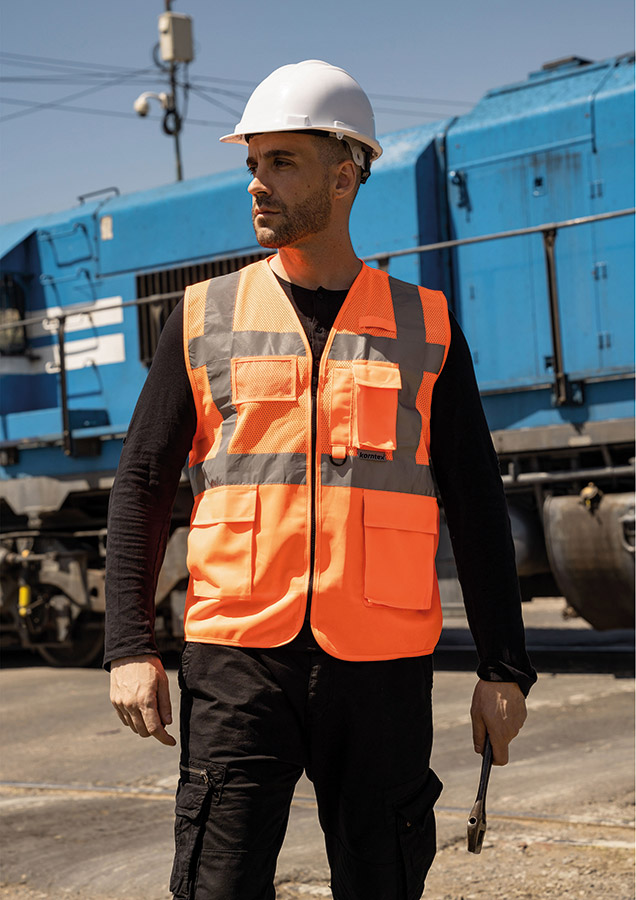 Adult worker with orange Korntex mesh multifunctional safety vest and helmet holding a tool on a railway site in front of a train