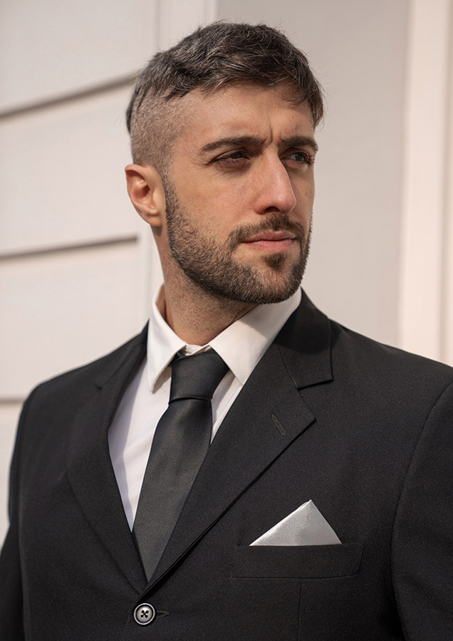 Elegant adult man in black suit with Korntex classic-tie and pocket handkerchief standing in front of a light-coloured wall