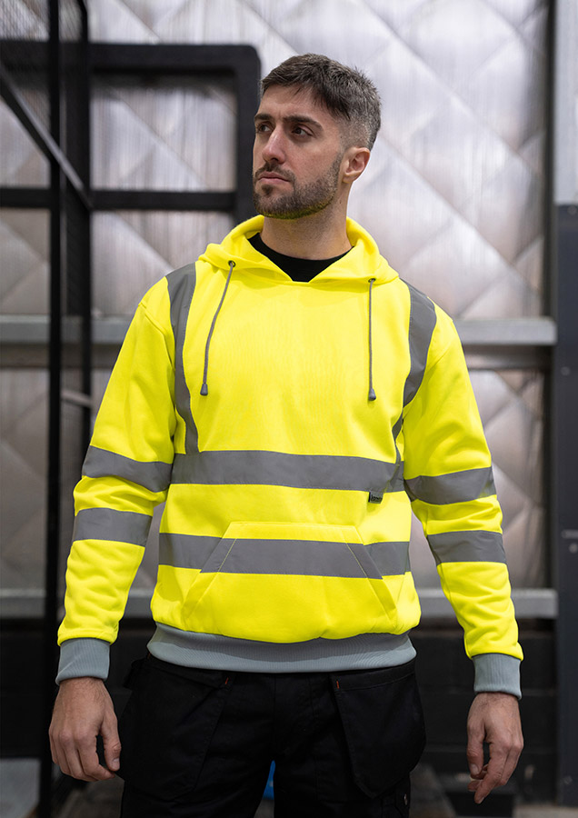 Adult man stands in signal yellow Korntex hi-vis hoodie with reflective stripes and black work trousers in an industrial environment