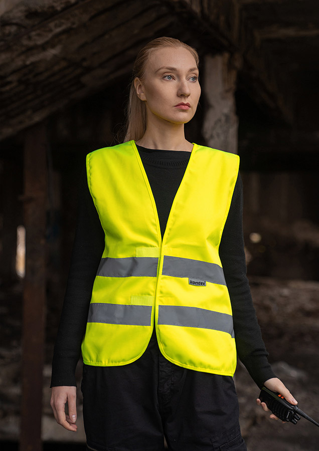 Adult woman in signal yellow Korntex safety vest with reflective stripes holding a radio in an industrial environment